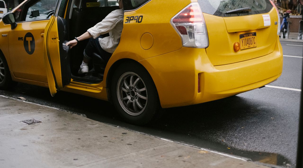 Contact Unrecognizable passenger closing door of yellow cab with logo and headlight parked on asphalt road near pavement on street in city